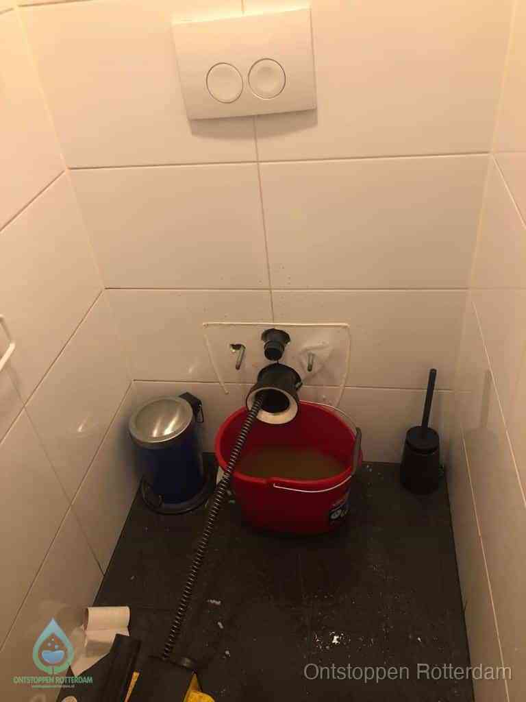 Een ontstopte wand-wc met verwijderde toiletpot waar een ontstoppingsveer in de wanduitgang verdwijnt, het achterhalen waar de verstopping zit in Rotterdam