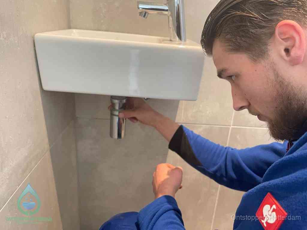 Een monteur draait met de hand de zwanenhalssifon onder een fonteinwasbak in de toiletruimte los om de bron van rioollucht in Rotterdam te vinden