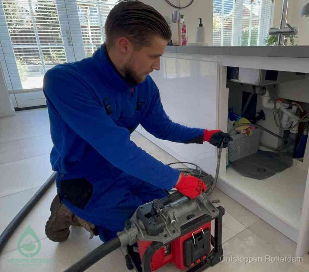 Een monteur met rode handschoenen knielt in een keuken en voert een elektrische ontstoppingsveer in tijdens het professioneel afvoer ontstoppen in Rotterdam
